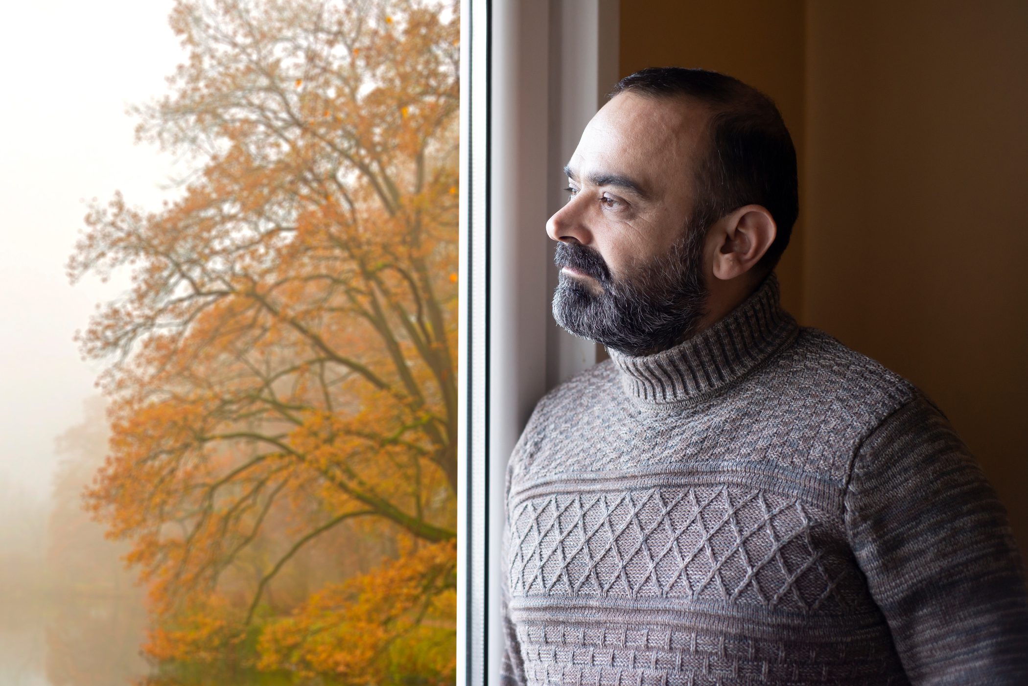 Man in a turtleneck sweater looks out a window on a foggy day.